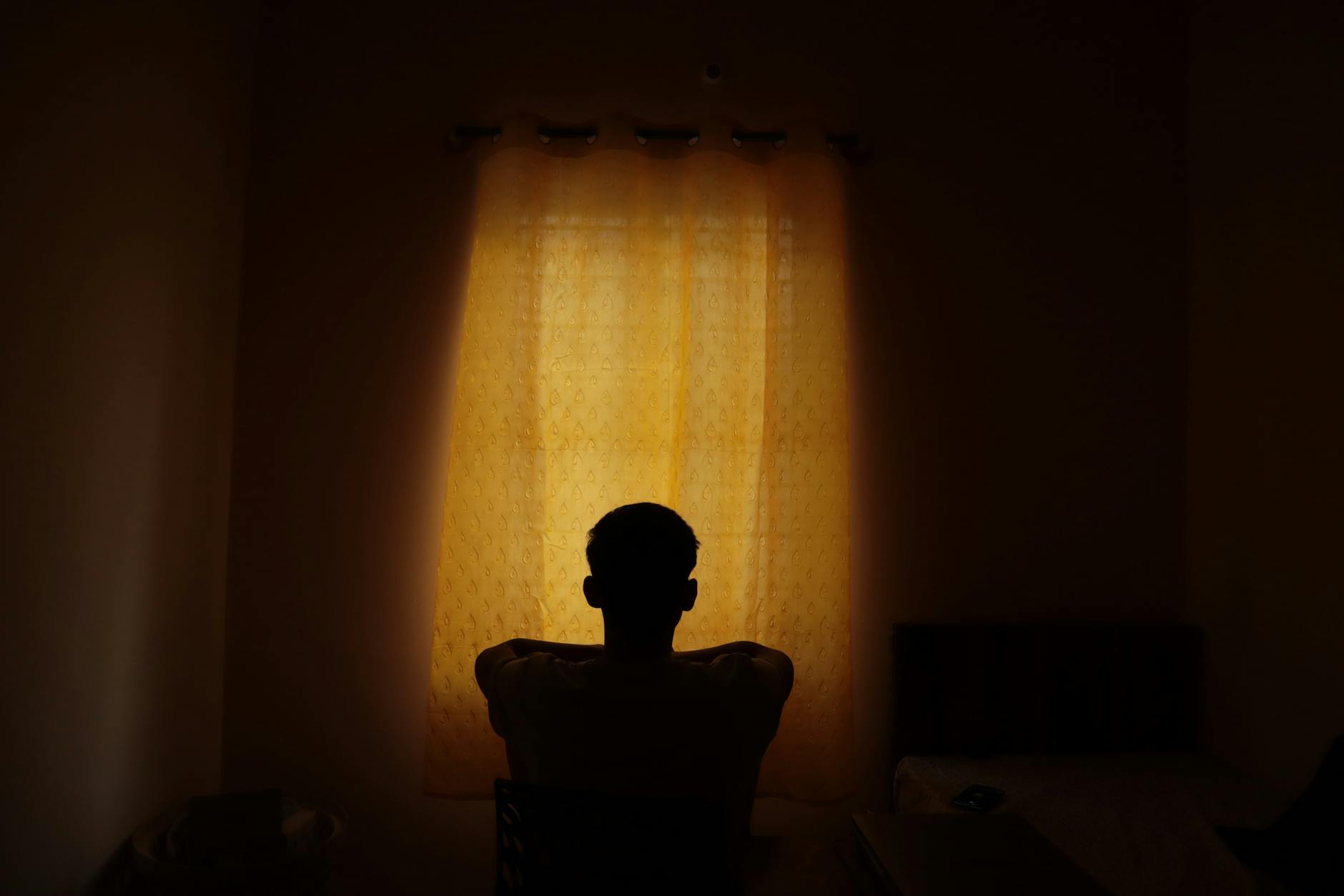 Eine Person sitzt im Halbdunkel vor einem Fenster, durch das warmes Licht fällt.